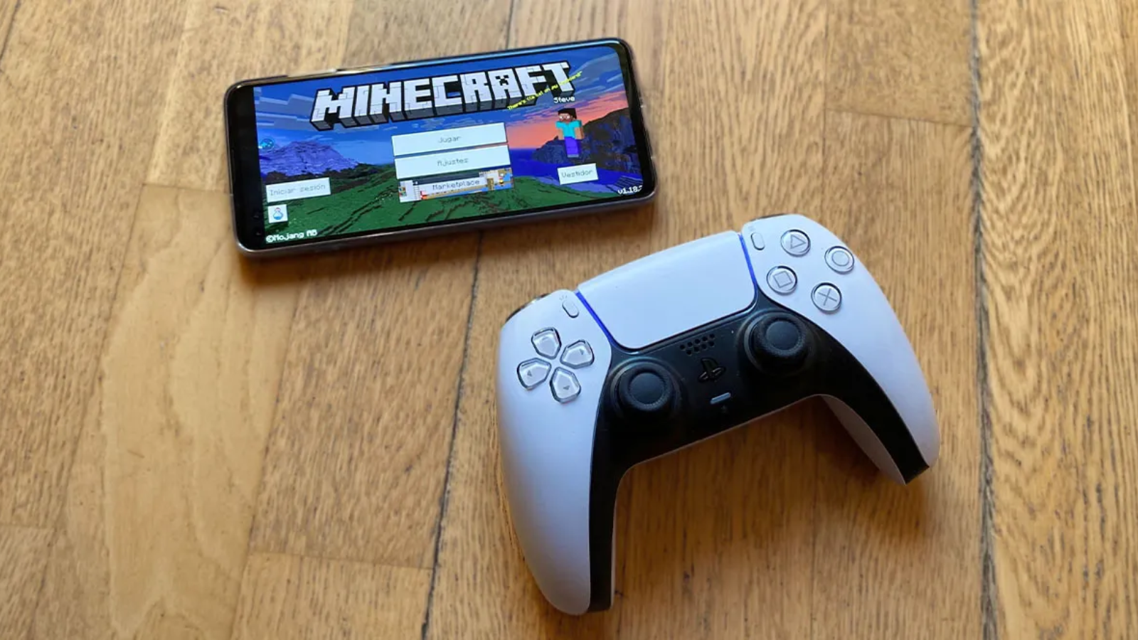 Mando DualSense de PS5 junto a un teléfono móvil que muestra la pantalla de inicio de Minecraft sobre un suelo de parqué.