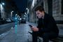 Hombre sentado en un banco de una calle nocturna consultando su teléfono iPhone con gesto de concentración.