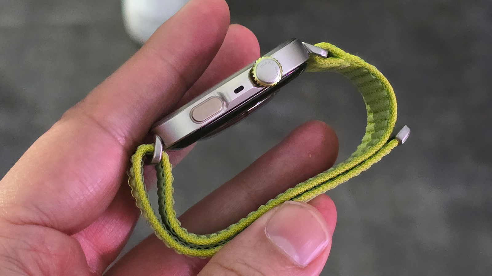 Primer plano lateral de un reloj inteligente con corona giratoria, botón físico y orificio de sensor, sostenido con los dedos.