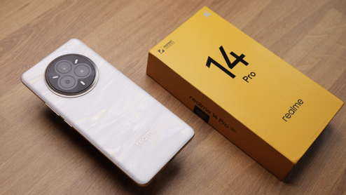 Parte trasera del realme 14 Pro en color blanco junto a su caja de venta amarilla sobre una mesa de madera.