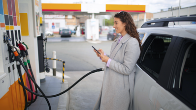 Mujer con abrigo gris utiliza un smartphone mientras llena el depósito de combustible de su coche en una gasolinera.