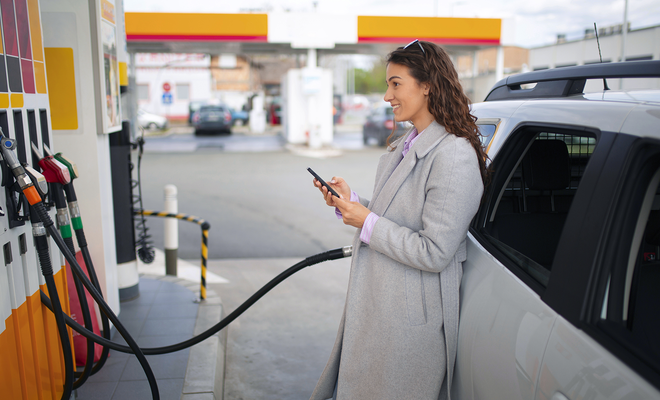 Mujer con abrigo gris utiliza un smartphone mientras llena el depósito de combustible de su coche en una gasolinera.