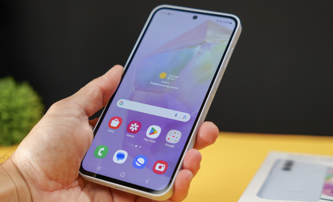 Mano sujetando un Samsung Galaxy A35 encendido mostrando la pantalla de inicio con iconos de aplicaciones y widget de clima.