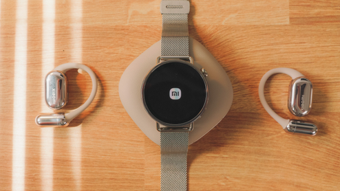 Reloj inteligente Xiaomi Watch S4 mostrando el logotipo de la marca en pantalla junto a unos auriculares de conducción ósea sobre una mesa de madera.