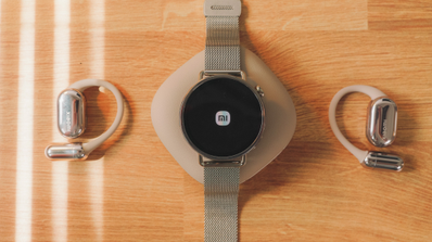 Reloj inteligente Xiaomi Watch S4 mostrando el logotipo de la marca en pantalla junto a unos auriculares de conducción ósea sobre una mesa de madera.