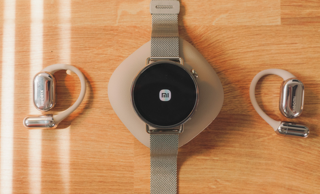 Reloj inteligente Xiaomi Watch S4 mostrando el logotipo de la marca en pantalla junto a unos auriculares de conducción ósea sobre una mesa de madera.