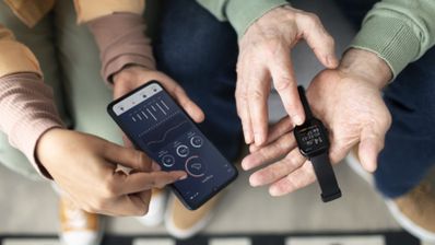 Primer plano de una persona mayor sujetando un reloj inteligente mientras otra maneja una aplicación de salud en un teléfono móvil.