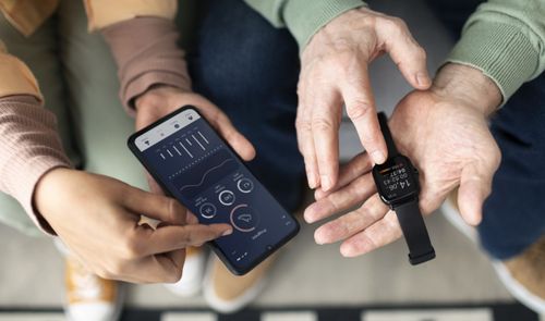 Primer plano de una persona mayor sujetando un reloj inteligente mientras otra maneja una aplicación de salud en un teléfono móvil.