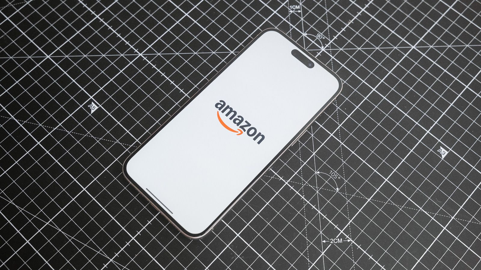 Logotipo de la aplicación de Amazon mostrado en la pantalla de un iPhone 16 Pro colocado sobre un tapete de corte negro.