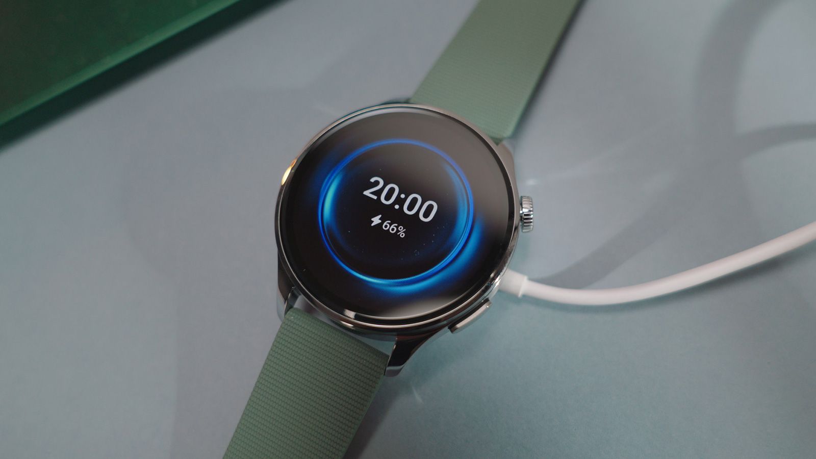 Reloj inteligente Xiaomi Watch 5 con correa verde sobre una superficie plana conectado a su cable de carga magnético.