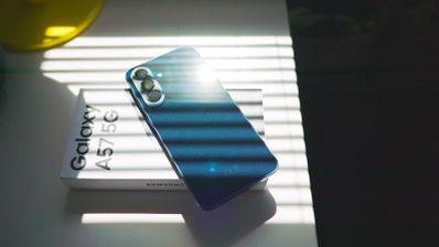 Parte trasera de un smartphone Samsung Galaxy A57 azul sobre su caja blanca, iluminado por la luz del sol a través de una persiana.