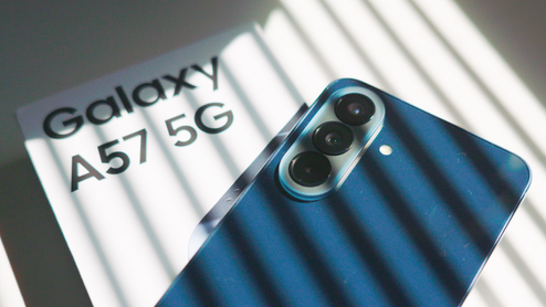 Parte trasera de un teléfono móvil de color azul que muestra sus tres cámaras junto a su caja con el texto Galaxy A57 5G bajo un juego de luces y sombras.