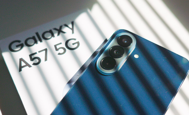 Parte trasera de un teléfono móvil de color azul que muestra sus tres cámaras junto a su caja con el texto Galaxy A57 5G bajo un juego de luces y sombras.