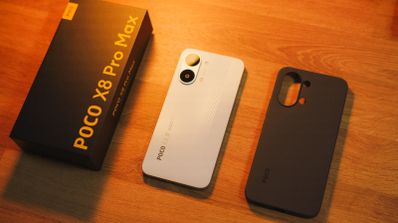 Parte trasera del teléfono POCO X8 Pro Max en color blanco junto a su caja y una funda protectora negra sobre una mesa de madera.
