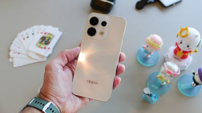 Mano sujetando un smartphone OPPO en color crema con un módulo de cámaras circular sobre una mesa con figuras decorativas y cartas.