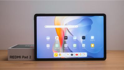 Pantalla frontal de la tableta Redmi Pad 2 encendida mostrando la interfaz de usuario con iconos de aplicaciones y barra de búsqueda sobre una mesa de madera.