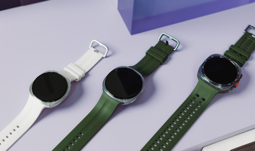 Tres relojes inteligentes Samsung Galaxy Watch alineados en diagonal sobre una superficie violeta, mostrando correas en color blanco y verde oliva.