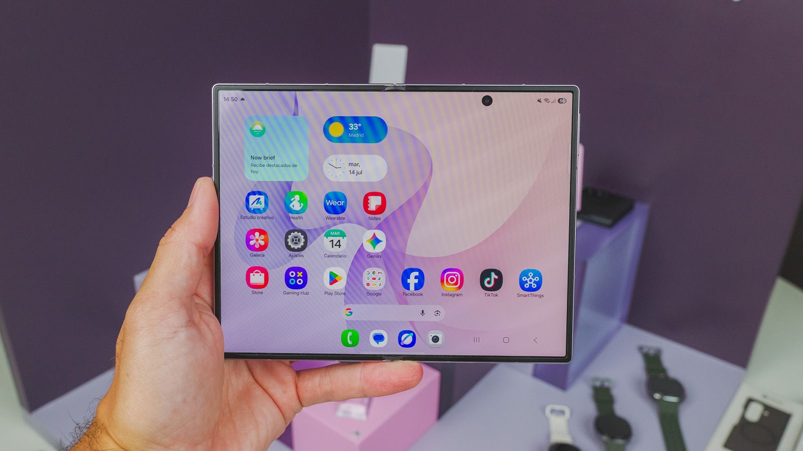 Mano sujetando un Samsung Galaxy Z Fold8 abierto mostrando la pantalla principal con iconos de aplicaciones y widgets.