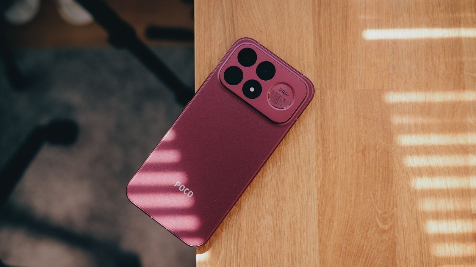 Parte trasera de un teléfono POCO F9 Ultra en color rojo cherry apoyado sobre una mesa de madera con sombras de persiana.