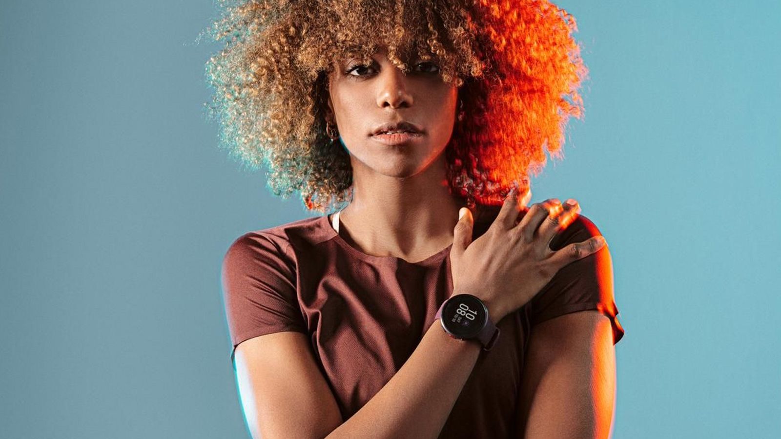 Mujer con pelo rizado posando con un reloj inteligente Polar en su muñeca izquierda sobre un fondo iluminado en azul y naranja.