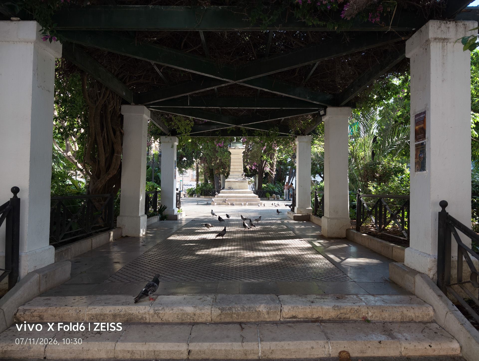 Perspectiva desde el interior de una pérgola hacia una plaza soleada con palomas, tomada con un vivo X Fold6.