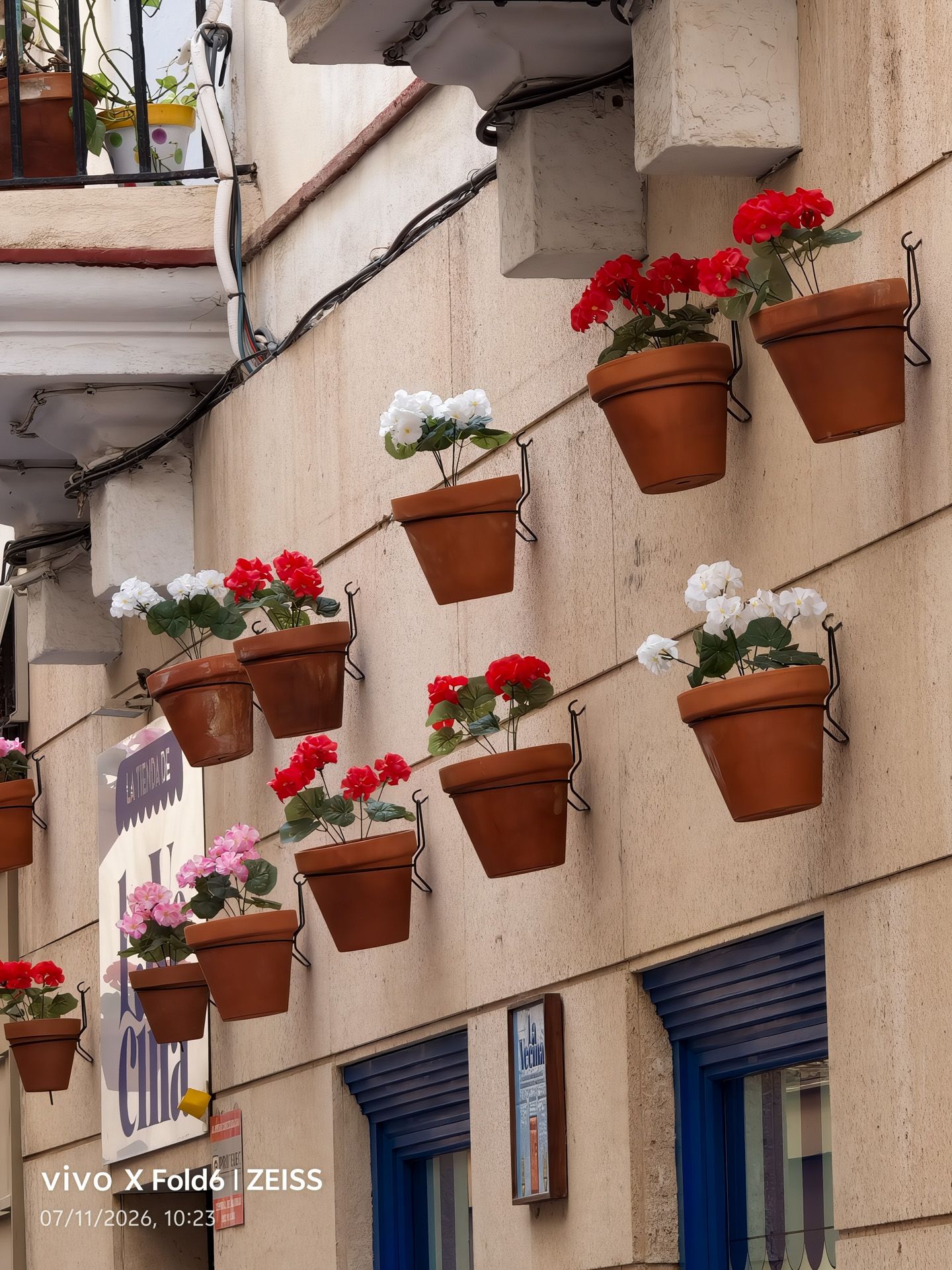 Fachada de un edificio decorada con macetas de barro colgantes y flores de colores, capturada con la cámara de un vivo X Fold6.