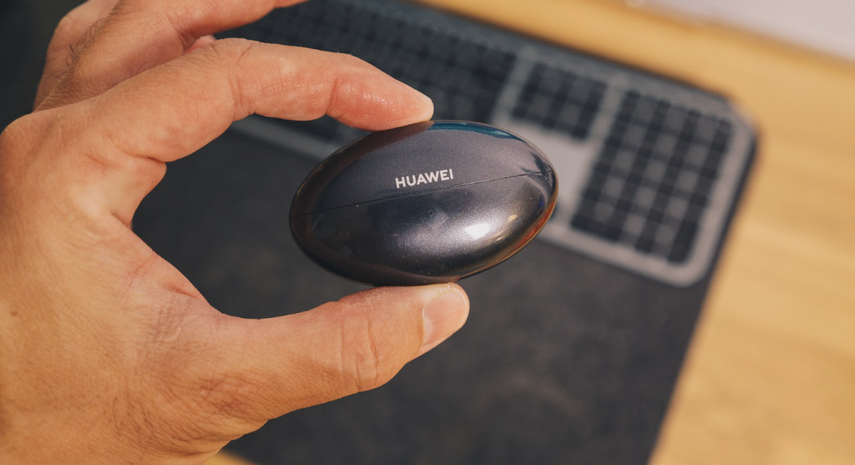 Primer plano del estuche de carga ovalado de los Huawei FreeBuds Neo sostenido con una mano sobre un escritorio.