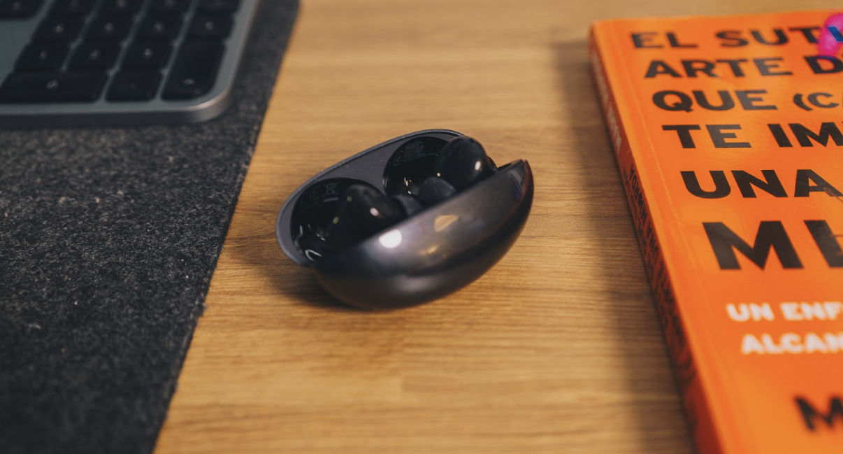 Estuche de carga de los Huawei FreeBuds Neo abierto sobre una mesa de madera junto a un libro naranja.