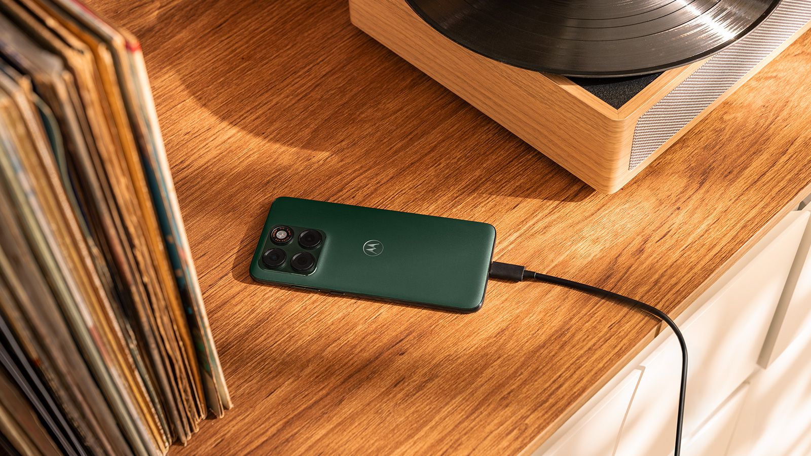 Motorola Edge 70 Plus de color verde oscuro conectado a un cable USB-C sobre un mueble de madera, junto a una colección de discos de vinilo y un tocadiscos.