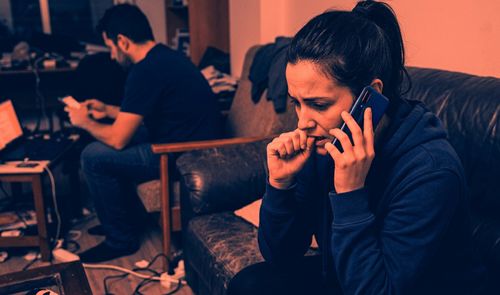Mujer con expresión de preocupación escuchando un audio de WhatsApp en su teléfono móvil mientras un hombre está al fondo en un salón.