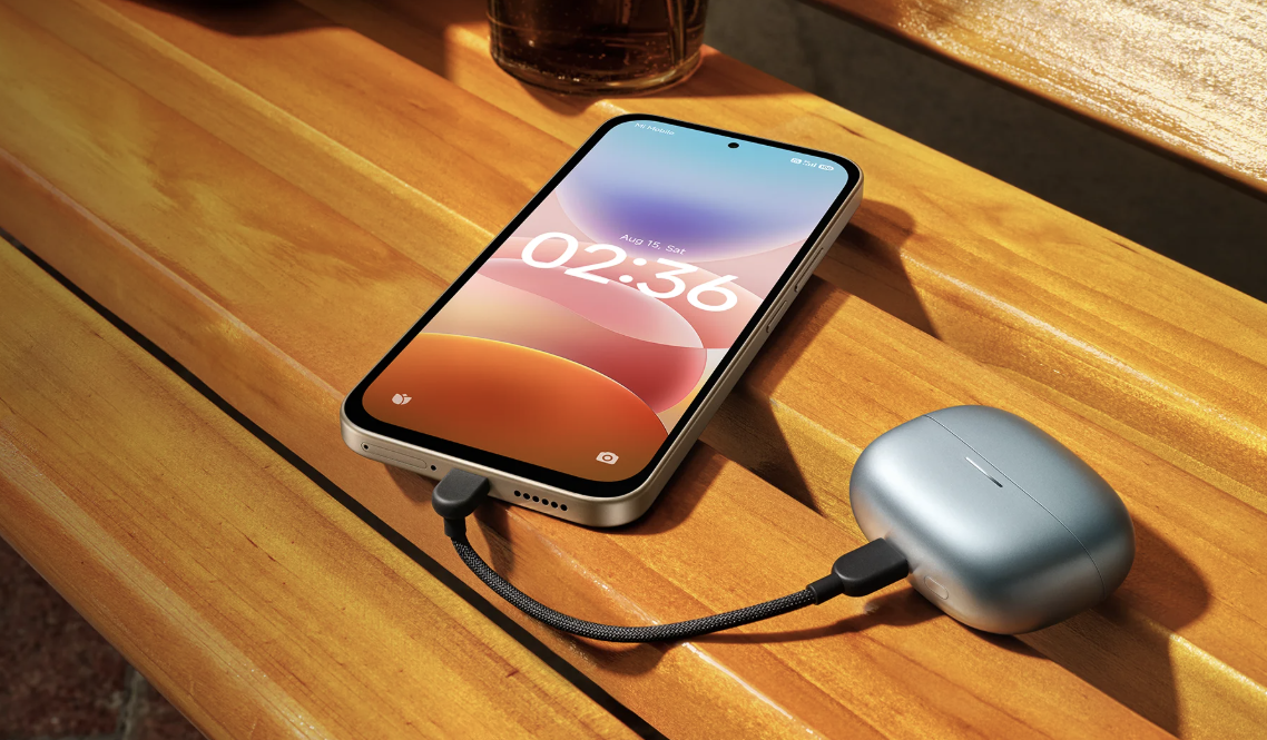 Teléfono Redmi 17 5G conectado por cable USB-C al estuche de unos auriculares inalámbricos sobre una mesa de madera.
