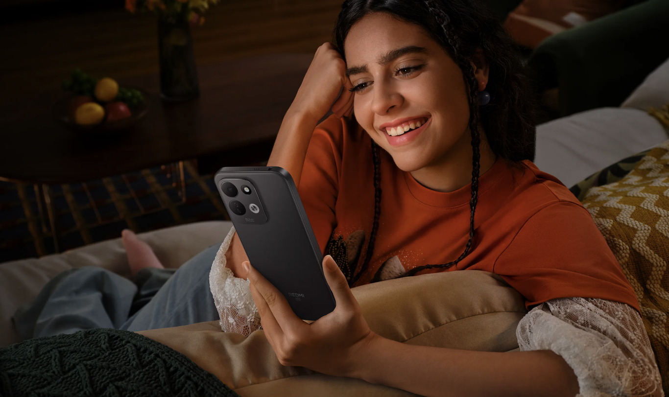 Una mujer joven sonríe mientras observa la pantalla de un smartphone Redmi 17 5G de color oscuro sentada en un sofá.