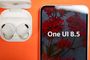 Primer plano de un Samsung Galaxy A55 mostrando el texto One UI 8.5 en pantalla junto a unos auriculares Galaxy Buds en su estuche sobre fondo naranja.