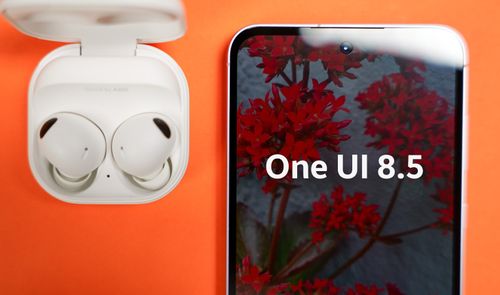 Primer plano de un Samsung Galaxy A55 mostrando el texto One UI 8.5 en pantalla junto a unos auriculares Galaxy Buds en su estuche sobre fondo naranja.