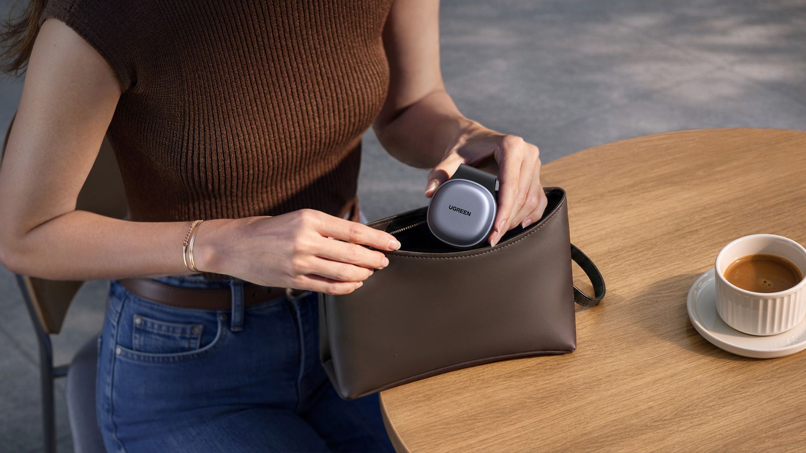 Una persona introduce el cargador inalámbrico plegable UGREEN MagFlow en un bolso pequeño sobre una mesa de madera.