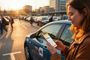 Mujer consultando una aplicación de alquiler de coches en su móvil junto a un vehículo en un aparcamiento al atardecer.