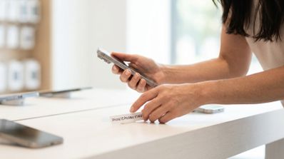Primer plano de una persona sosteniendo un iPhone y revisando una etiqueta de precio en el mostrador de una tienda oficial.