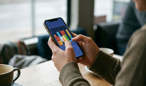 Persona jugando a un videojuego de rompecabezas con bloques de colores en un smartphone mientras toma café.