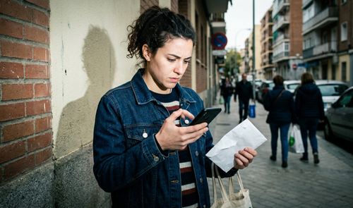 Mujer joven con gesto de preocupación mirando su teléfono móvil mientras sostiene documentos en una calle de la ciudad.
