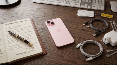 iPhone 15 Plus en color rosa sobre una mesa de madera junto a cables de carga y una libreta abierta.