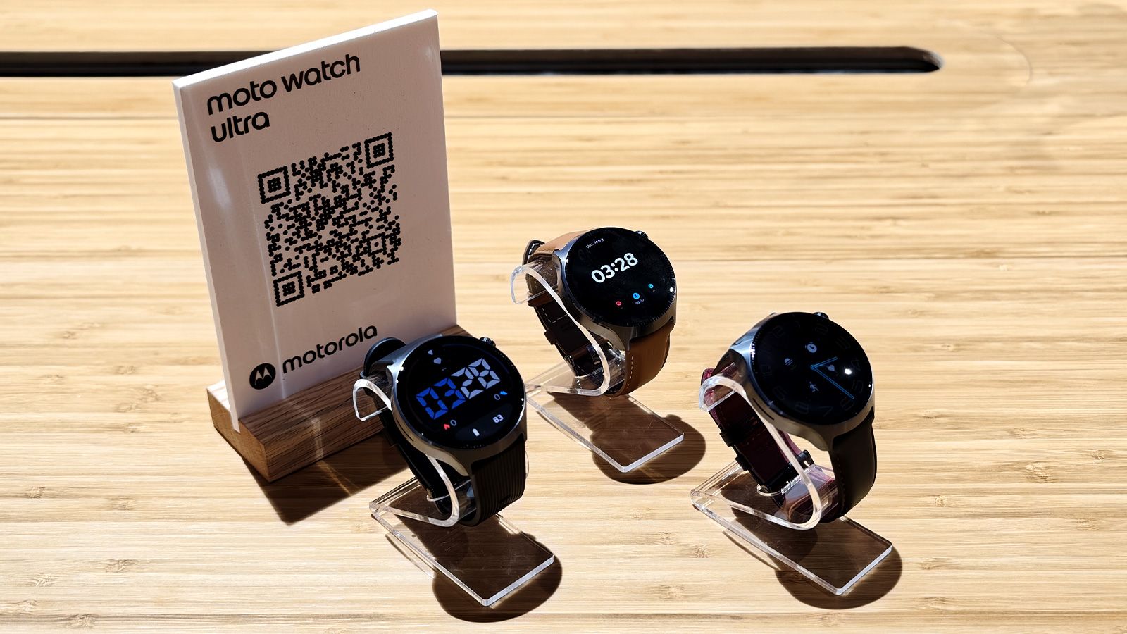 Tres relojes inteligentes Moto Watch Ultra expuestos en soportes transparentes junto a un panel con código QR en la feria IFA.