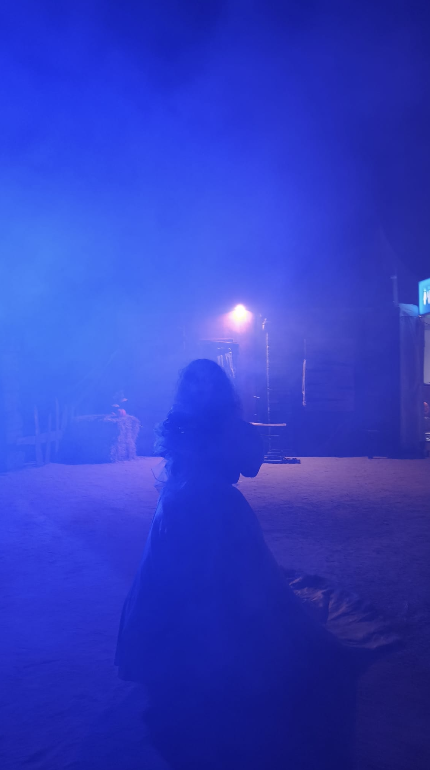 Silueta de una persona con pelo largo bajo una densa niebla azul en un escenario nocturno con iluminación artificial.
