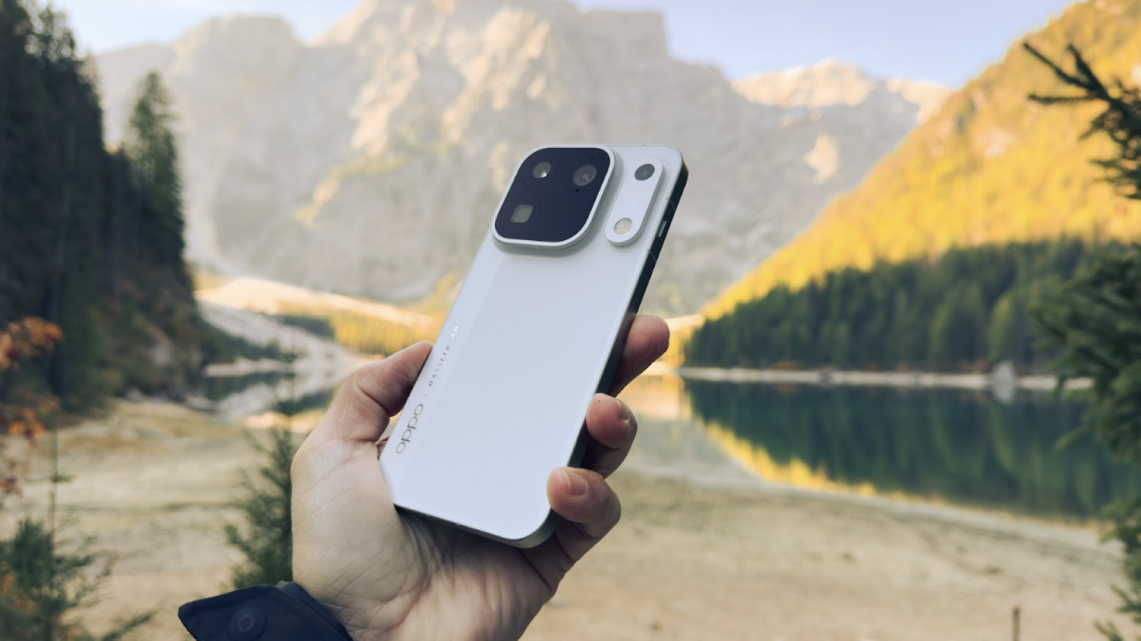 Mano sujetando el teléfono móvil OPPO Find X10 Pro Max en color blanco frente a un lago de montaña y picos rocosos.