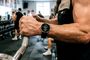 Primer plano de un hombre utilizando un reloj inteligente mientras sujeta una barra de pesas en un gimnasio.