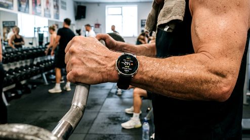 Primer plano de un hombre utilizando un reloj inteligente mientras sujeta una barra de pesas en un gimnasio.