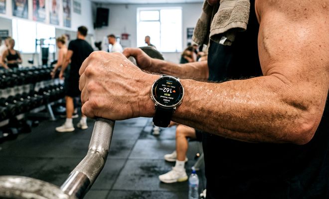 Primer plano de un hombre utilizando un reloj inteligente mientras sujeta una barra de pesas en un gimnasio.