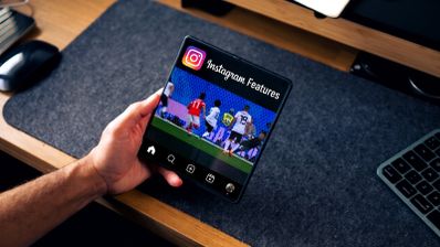 Mano sujetando un teléfono plegable abierto que muestra la interfaz de Instagram sobre un escritorio.