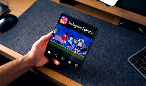 Mano sujetando un teléfono plegable abierto que muestra la interfaz de Instagram sobre un escritorio.