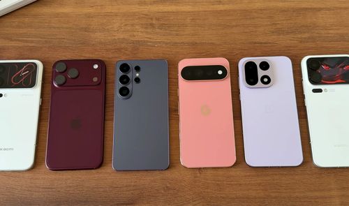 Seis smartphones de diferentes marcas y colores colocados en fila sobre una superficie de madera, mostrando sus módulos de cámara traseros.