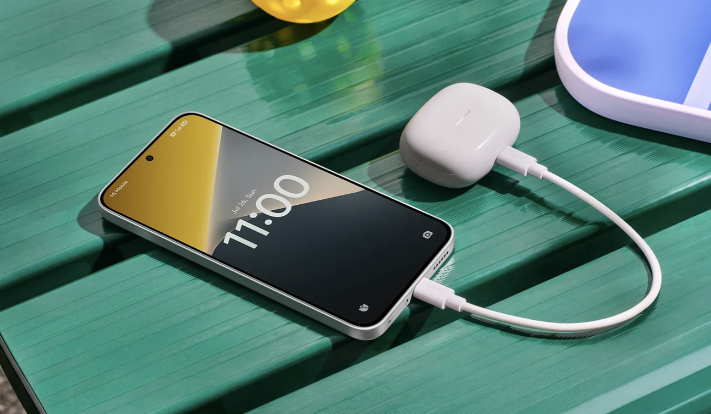 Smartphone POCO X8 conectado mediante cable USB-C a un estuche de auriculares inalámbricos sobre una mesa verde.
